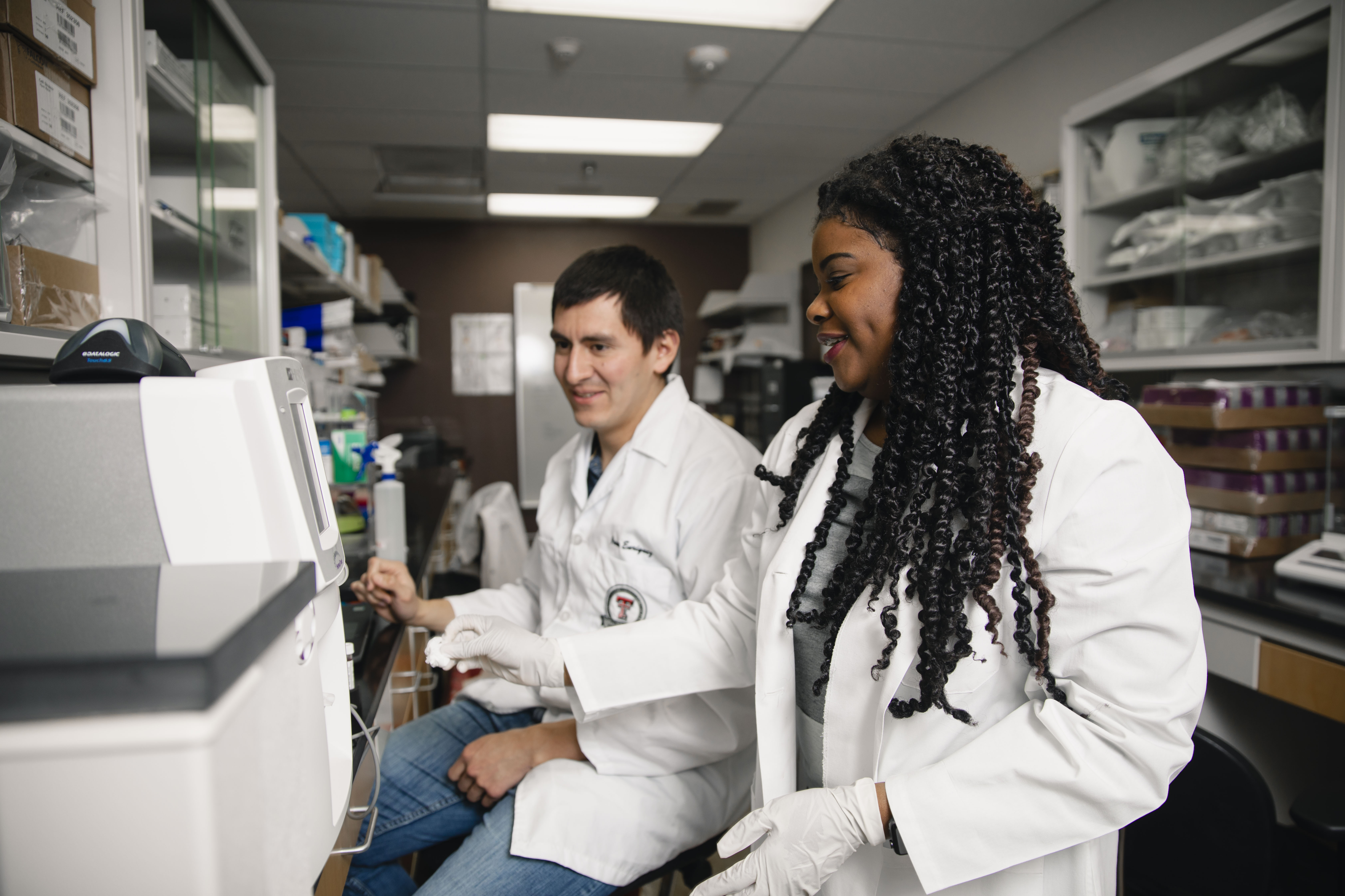 A male and female student in white coats working in lab