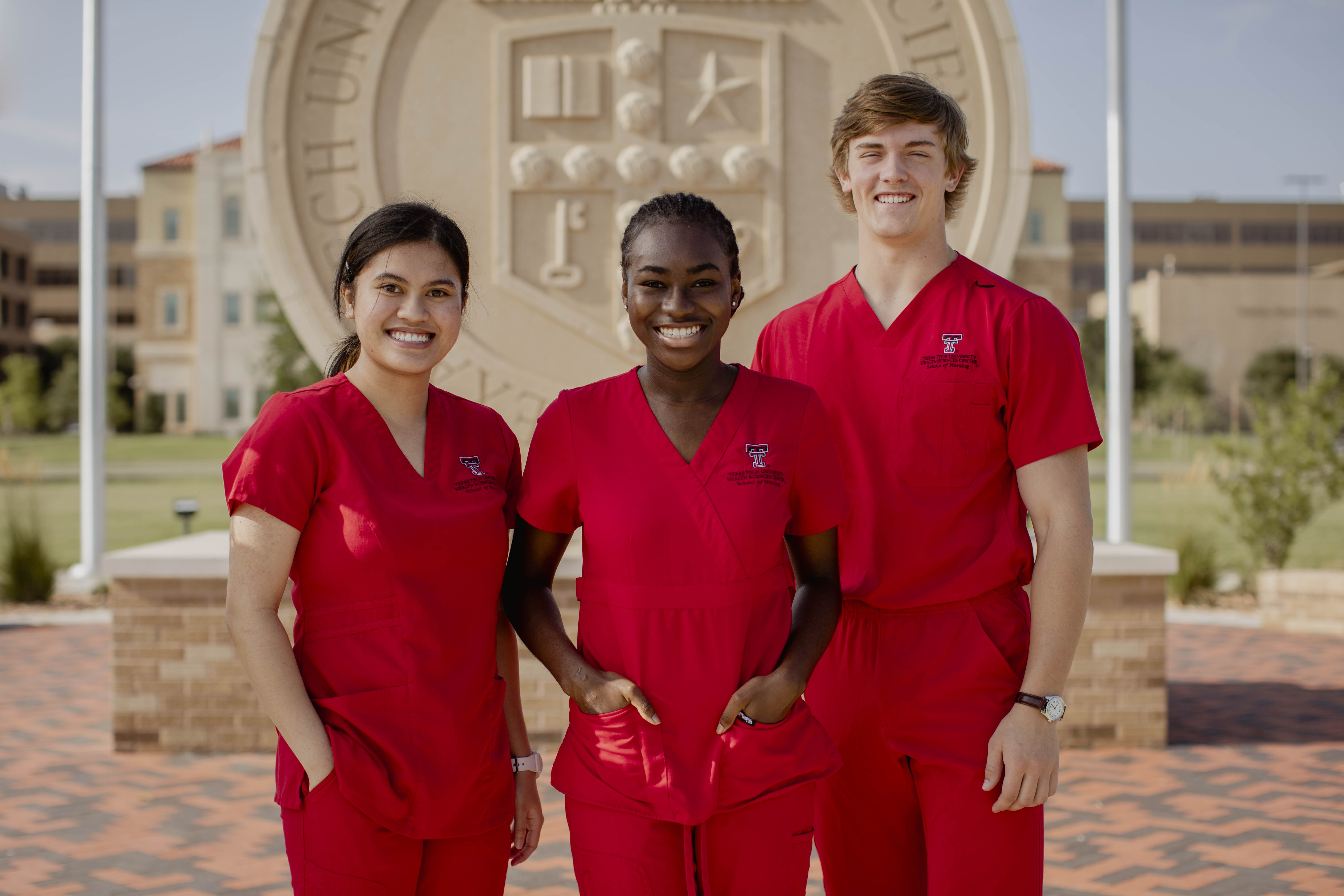 three nursing students smiling in front of University seal in red scrubs